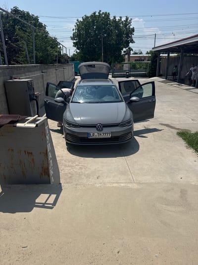 At a self-service car wash in Belozem, Bulgaria, a gray Volkswagen Golf 8 wagon with an open trunk and front passenger door is parked on a concrete surface under a bright, sunny sky. A person, wearing a light-colored shirt and dark shorts, is bent over reaching into the car through the open passenger door. To the left of the car, a rusty, industrial-style dumpster with white lettering, including "ТУПАЛКА", is positioned near a gray metal cabinet. In the background, a covered structure, likely part of the car wash facility, is visible, with two men standing near it. Trees and buildings are also present in the background, indicating an outdoor urban or semi-urban environment. Power lines crisscross the sky above.