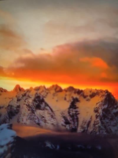 A panoramic view showcases snow-capped mountains against a vibrant sky. The upper portion is dominated by an orange and peach-hued sky, filled with wispy clouds. Below, the jagged peaks of the mountains are partially covered in snow, casting shadows on their rocky faces. The foreground features a darker, undefined area, possibly representing lower terrain or bodies of water. The overall scene conveys a sense of grandeur and natural beauty, emphasizing the contrast between the warm sky and the cool mountains.