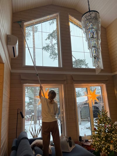 Inside a residence in Finskiy Briz, Russia, a blonde-haired individual, seen from behind, is actively cleaning a tall interior window. The person, wearing a light-colored short-sleeved top and dark trousers, stands on a dark blue sofa and extends a long, telescopic pole with a green cleaning head towards the upper portion of a large, multi-pane window. The room features light wood-paneled walls and a high, sloped ceiling. An air conditioning unit is mounted on the left wall. To the right, a multi-tiered crystal chandelier hangs prominently. Below the large upper windows, two lower windows are adorned with large, illuminated star-shaped decorations. A fully decorated Christmas tree, illuminated with lights, stands to the right. The exterior view through the windows reveals a snow-covered landscape with pine trees and what appears to be another snow-dusted structure or gazebo, along with outdoor string lights. A black floor lamp and a grey cat-shaped pet bed are visible in the foreground.
