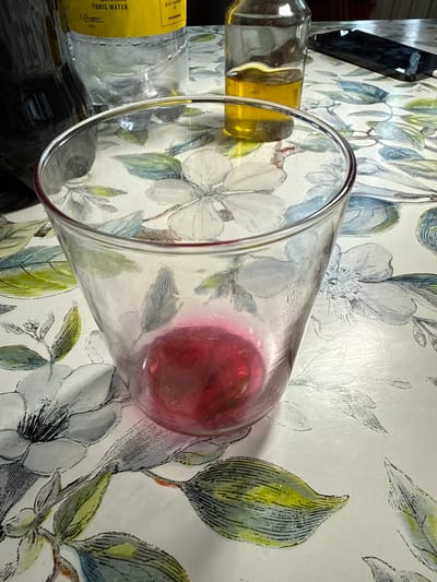 The image shows a close-up, angled view of a clear glass tumbler containing a red liquid. The tumbler is positioned on a surface with a floral patterned wallpaper, featuring stylized depictions of white flowers and green leaves. To the left of the tumbler, partially visible, is a clear glass bottle with a yellow label that reads "ORIGINAL TONIC WATER". Behind the tonic water bottle, another bottle with a yellow liquid is visible. The background wallpaper has a vintage aesthetic with botanical illustrations in muted colors. The lighting suggests an indoor setting, possibly a kitchen or dining area, with no indication of the time of day or weather. The scene is static, with no people or active events depicted. The focus is on the objects and their arrangement on the patterned surface.