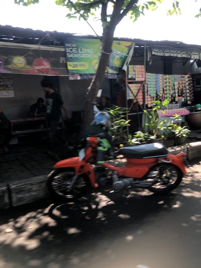 This image depicts an outdoor scene in Buduran, Indonesia, likely a roadside stall or small shop. In the foreground, an orange motorcycle is parked, its front wheel and part of the engine visible. The motorcycle casts a shadow on the wet pavement, which reflects dappled sunlight filtering through overhead trees. Behind the motorcycle, a stall is set up, with a corrugated metal roof supported by wooden posts. A banner advertising "NEW ICE LIME SENSATION" is prominently displayed, along with prices in Rupiah (Rp 26,500). Various products, likely snacks or drinks, are neatly arranged on shelves or hung from the stall's structure. Two individuals are partially visible within the stall area, one appearing to be walking and the other seated. The overall atmosphere suggests a commercial area in a tropical climate, with evidence of a recent rain shower due to the wet pavement and reflections.