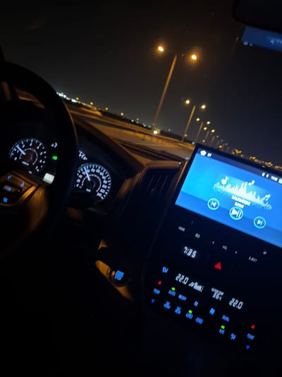 The image captures the interior of a car at night, viewed from the driver's or front passenger's perspective, while driving or stationary on a road in Umm Salal Ali, Qatar. The scene is dominated by the illuminated car dashboard and the dark exterior with distant streetlights.

In the center, a large infotainment screen displays a music player interface, showing an equalizer graphic and the text "Kavalan GAYAN," alongside "HD" and control icons. Below the screen, the climate control panel is visible with various illuminated buttons and displays. Notable readings include the time "7:39," an interior temperature of "22.0" degrees Celsius, and an "OUTSIDE TEMP" of "22.0" degrees Celsius. Buttons for "TEMP," "DUAL," "A/C," and "AUTO OFF" are clearly visible, glowing in red, blue, and green.

To the bottom left, part of the steering wheel is visible, and behind it, the car's instrument cluster with two glowing circular gauges. One gauge, likely the speedometer, shows markings up to "240," with specific numbers like "100," "120," "140," "160," "180," "200," "220," and "240" clearly readable.

Outside the car, a long line of warm-toned streetlights stretches into the dark night, suggesting a highway or a main road. The overall ambiance is dark and calm, characteristic of a night drive.