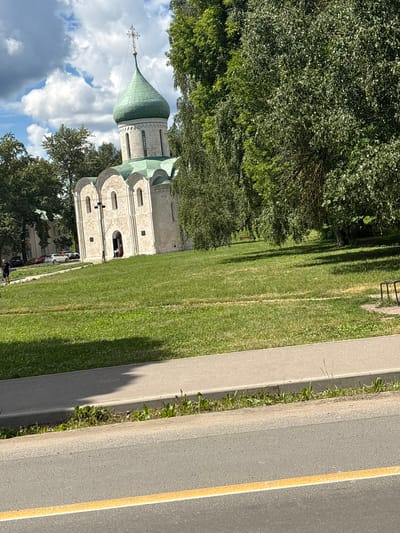 The image shows the Church of St. Theodore the Black in Pereslavl-Zalessky, Russia, on a sunny day. The white stone church has a green, onion-shaped dome topped with a cross. It is surrounded by a well-maintained green lawn and mature trees. A paved sidewalk runs along the foreground, with a road beyond it. In the background, several cars are parked near another building. A person in dark clothing is walking on the lawn. The sky is blue with scattered white clouds.