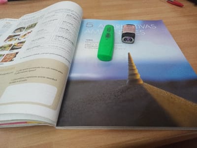 The scene depicts an open textbook or magazine resting on a light-colored wooden surface. The specific location within San Marzano sul Sarno, Italy, is not discernible from the image. No individuals are present.
The open publication spans the central area of the frame. The left page displays text, primarily in Spanish, with sections related to grammar and vocabulary, accompanied by small photographic insets of various food dishes, including pizza. The right page features a large photograph depicting a sandy, arid landscape under a light blue sky, with a prominent, spiraling conical sand dune or structure casting a long shadow across the foreground. Overlaid on this image are the words "5 ALTERNATIVAS AMBIENTALES".
Two stationery items are placed on the right page of the book: a bright green cylindrical highlighter pen with a white cap, positioned horizontally, and a small, black, and pink rectangular prism-shaped pencil sharpener. The sharpener features a white circular design resembling a panda's face on one end.
The wooden surface beneath the book exhibits a distinct grain pattern. Partially visible at the extreme top left of the frame is the tip of a black writing instrument. Similarly, the red tip and a segment of a red writing instrument are partially visible at the top right of the frame. The objects are static, arranged as if in a study or work setting.