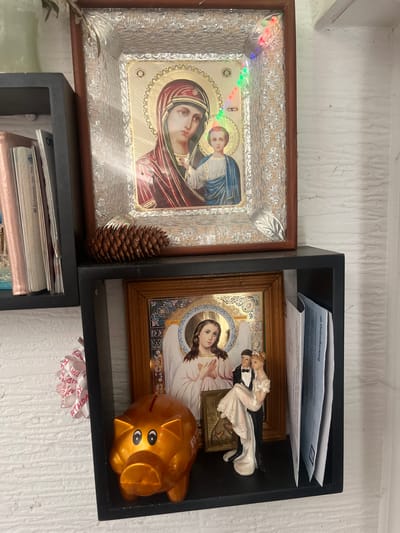 The image shows a close-up of a shelf in what appears to be a home in Schwäbisch Hall, Germany. The shelf is filled with religious and decorative items. On the top shelf, there is a framed icon of the Virgin Mary holding the infant Jesus, with a pinecone placed in front of it. To the left of the icon, a stack of books is visible. The bottom shelf contains another framed religious image, this one depicting an angel. Next to the angel image is a gold piggy bank and a small figurine of a bride and groom, possibly a cake topper. To the right of these items are several folded papers, some appearing to be mail or documents. The background is a textured white wall, consistent with common interior finishes. The overall impression is of a personal collection of items displayed on a shelf in a domestic setting.