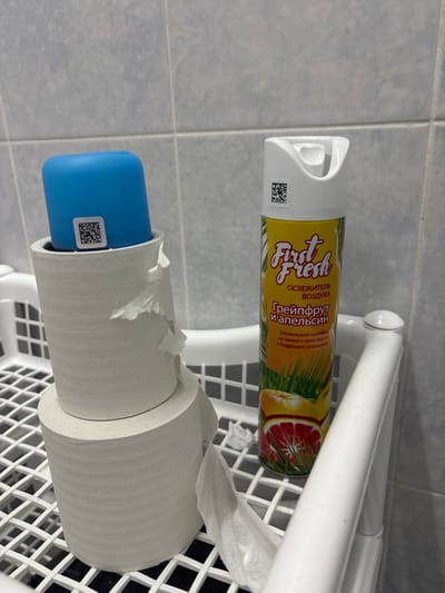 The image shows two bathroom items on a white plastic shelf against a light blue tiled wall. The top item is a white aerosol can of air freshener with a yellow label featuring a graphic of grapefruit and palm leaves. The label has text in Russian, including "First Fresh," "ОСВЕЖИТЕЛЬ ВОЗДУХА" (Air Freshener), "Грейпфрут и апельсин" (Grapefruit and Orange), and a description of the scent as a "Tropical cocktail of fresh grapefruit and invigorating orange." There is a small QR code on the side of the can.
Below the air freshener is a roll of toilet paper. A blue plastic dispenser is inserted into the core of the toilet paper roll, and this dispenser also has a small QR code on its side. The toilet paper is slightly unraveled, with loose sheets hanging off the roll. The overall scene suggests a typical bathroom setting in Novocherkassk, Russia. The lighting appears to be artificial, typical of an indoor environment. There are no people visible in the image.