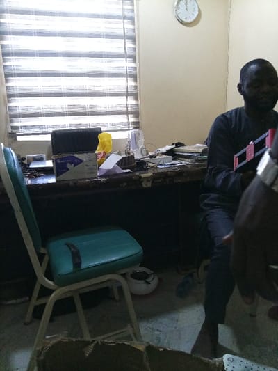 This image depicts an indoor scene in Gwammaja, Nigeria, characterized as an office or workspace. The room features light-colored walls and a window on the left covered by horizontal venetian blinds with alternating sheer and opaque strips. A circular wall clock displaying Arabic numerals is mounted on the upper right wall, indicating approximately 08:30. The floor appears to be concrete or tiled.

In the mid-ground, a dark-colored wooden desk, showing significant wear with chipped paint or veneer, is positioned. The desk is cluttered with items including a "Smart Care" branded box featuring blue gloves, a yellow plastic bag, stacks of papers, and various electronic components and wires. A small transparent plastic container with a vertical antenna-like element is also present on the desk. A laptop or similar device is partially visible to the left of the glove box.

An adult male, with dark skin, short dark hair, and a beard, is seated at the desk on the right side of the frame. He wears a dark long-sleeved shirt and dark trousers, and his bare feet are visible under the desk. His face is slightly blurred and directed to the right, out of frame. He is holding a black and red rectangular object, possibly an electronic device or tool, featuring multiple horizontal red bars.

To the left of the desk, a light-framed chair with a teal/aqua padded seat and backrest is positioned. A dark, curved object rests on the chair's seat. Under this chair, a white ceramic object with a red base, resembling an ashtray or bowl, is visible on the floor, alongside a blue plastic bottle beneath the desk. The immediate foreground is partially obscured by a torn cardboard box. A portion of another individual's arm and hand, adorned with a metallic wristband or watch, is visible on the extreme right foreground, indicating the presence of a second person.