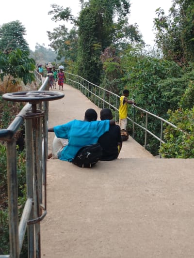 The image captures an outdoor scene featuring a concrete pathway or bridge winding through a densely vegetated, possibly tropical, environment under a bright, somewhat overcast or hazy sky. The perspective is slightly elevated, looking along the path.
In the immediate foreground on the left, a rusty metal structure, appearing to be a large handwheel or valve control, is prominently featured, attached to a metal railing. This railing extends along the left edge of the pathway, disappearing into the distance.
Midground, two individuals are seated on the concrete pathway, facing away from the camera and looking towards the right side of the frame. The person on the left is wearing a bright, light blue, loose-fitting garment, possibly a tunic, over light-colored trousers. Their right arm is extended, resting around the shoulders of the second person. This second individual wears a dark-colored top (likely black) and light-colored bottoms. A black backpack with white logos is positioned on the ground in front of the person in the dark top. Their posture suggests they are resting or observing their surroundings.
Slightly further along the path and to the right of the seated pair, a young child stands next to the metal railing, facing outwards from the path. The child is wearing a bright yellow sleeveless top and light-colored shorts. They appear to be holding a small, light-colored object in their right hand, possibly a cup or a bottle, and are looking out over the railing into the vegetation.
In the distance, along the curved pathway, several figures, primarily women, are visible walking towards the camera. Many of them are carrying items, such as baskets or bundles, balanced on their heads, a traditional method of carrying goods. Their clothing appears colorful and patterned. They are walking on the same concrete path, which is flanked by similar metal railings on both sides.
The background and surrounding areas are dominated by abundant, lush green foliage. Tall trees with dense canopies line both sides of the pathway, creating a vibrant natural setting. The pathway itself appears to be well-constructed, suggesting it might be part of an infrastructure project like a bridge or an elevated walk in a rural or natural landscape. The overall mood of the image is calm and depicts everyday life in a verdant setting. There is no discernible text within the image.