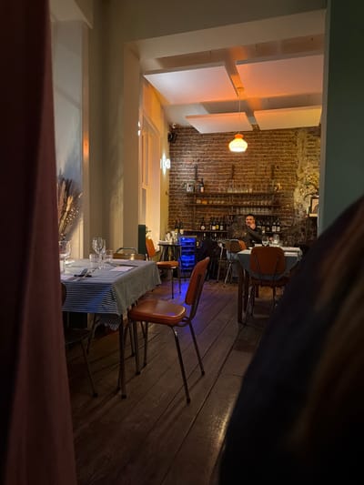 The image depicts the interior of a commercial establishment, identified as a restaurant or bar, located in Ixelles - Elsene, Belgium. The scene is illuminated by warm, artificial light sources, including a central pendant lamp with an orange-domed shade and various wall-mounted fixtures, notably a vertical white "10B" light sign. The background wall is exposed red brickwork, featuring a metal shelving unit functioning as a bar, stocked with multiple bottles of wine and spirits, alongside assorted glassware. A blue-illuminated beverage cooler is positioned on this bar setup.

The floor is comprised of dark wooden planks. Several dining tables are arranged across the room; those in the foreground and mid-ground are covered with blue and white checkered tablecloths, set with clear wine glasses, tumblers, and cutlery. Chairs with orange-brown upholstered seats and metallic frames are positioned around these tables.

One adult male with dark hair, wearing a dark long-sleeved garment, is seated at a table near the bar area, looking generally towards the left side of the frame with a subtle smile. Above, white rectangular acoustic panels are suspended from the ceiling. A security camera is mounted high on the brick wall. In the immediate left foreground, a blurred reddish-brown fabric, possibly a curtain, partially obscures the view. A dark, blurred object occupies a portion of the lower right foreground.