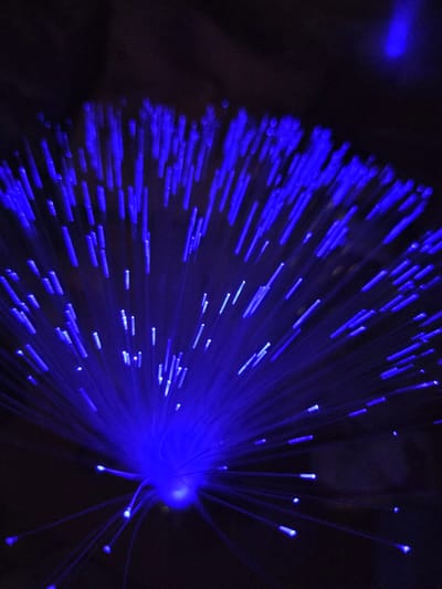 A close-up shot shows a fiber optic lamp emanating blue light. The light sources are arranged in a radial pattern from the base and are set against a dark background. No people are visible in the image. The location is not discernible from the visual information.