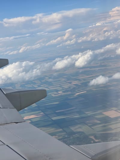 An aerial view from an airplane window shows a patchwork of agricultural fields and clouds. The wing of the airplane is visible in the foreground, taking up the lower left portion of the image. The fields below are a mix of green, brown, and yellow, indicating different crops or stages of growth. Some clouds are scattered across the sky, casting shadows on the landscape. A small village or cluster of buildings can be seen in the distance, nestled among the fields. There are no people or specific actions depicted in the image. The scene is peaceful and expansive, capturing a typical agricultural landscape from high above. The location is identified as Kremena, Bulgaria.