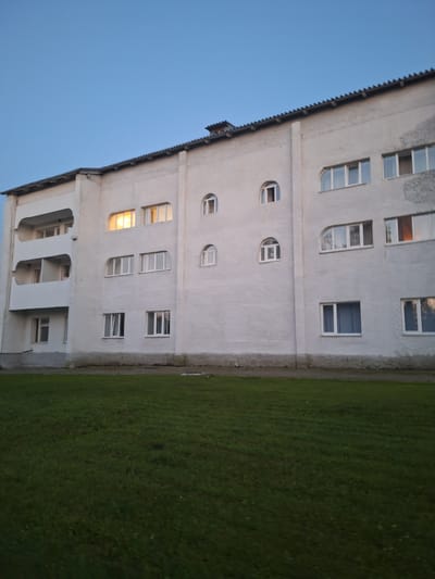A low-angle outdoor shot captures the exterior of a multi-story apartment building in Atamanovka, Russia, under a clear, pale blue twilight sky. The building, painted white and showing signs of weathering, features several rows of windows and balconies. On the left side, there are three levels of recessed balconies. Various window shapes are visible, including rectangular, arched, and semi-circular designs. One set of upper-floor windows on the left side is illuminated, indicating interior lights are on. The building stands adjacent to a well-maintained grassy area. The overall scene depicts a quiet residential setting.