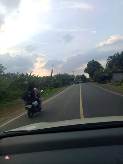 An over-the-shoulder shot captures a motorcycle traveling on a paved road in Halaban, Indonesia. The motorcycle is carrying two individuals, one driving and one as a passenger, both facing away from the camera. The passenger has a large black bag strapped to their back. Lush green vegetation, including what appears to be banana trees, lines the left side of the road. The right side of the road features a few simple structures, one of which has a weathered wooden exterior and a corrugated metal roof, and several palm trees. The road itself is asphalt with a yellow line marking the center. The sky above is overcast with dramatic clouds, suggesting either early morning or late afternoon light. The foreground of the image shows the dashboard and windshield of a vehicle, indicating the photo was taken from inside a car.