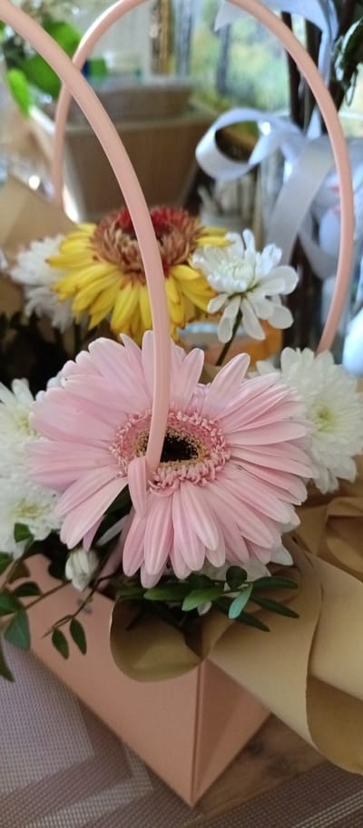 The image shows a close-up of a flower arrangement in a container with a handle, likely a gift or decorative item. A large pink gerbera daisy is the most prominent flower, with a dark center and numerous petals. Other flowers in the arrangement include white and yellow daisies, adding to the bouquet's diverse color palette. Green leaves are visible, providing a natural backdrop to the flowers. The container is peach colored with a beige ribbon. The background is blurry, suggesting an interior setting. The location is provided as Irbit, Russia. No people are visible in the image.
