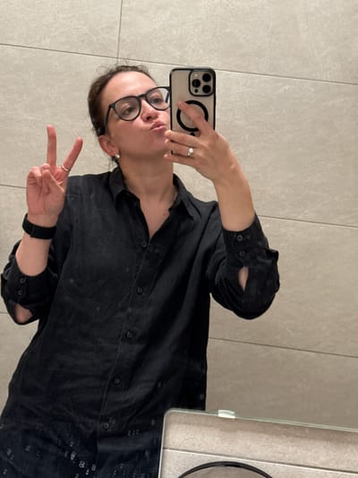 A person is taking a selfie in a bathroom with gray tile walls. The person has glasses and is wearing a black shirt and a black watch. They are making a peace sign with their left hand. The location is unspecified, and there is no information to indicate that the image was taken in Khyrkasy, Russia.
