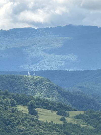 The image depicts a layered mountainous landscape. In the foreground, rolling green hills are covered with dense trees and a few patches of mown grass. Two tall, white telecommunication towers stand prominently on a hilltop in the middle ground, surrounded by trees. Behind these, successive ranges of mountains recede into the distance, their colors shifting from deep green to muted blues, indicating atmospheric perspective. The sky above is overcast with soft, white clouds. The scene is devoid of human presence, focusing solely on the natural environment and the man-made structures. This vista appears to be in or near Orbeli, Georgia, given the context provided, characterized by its verdant, hilly terrain and atmospheric conditions.