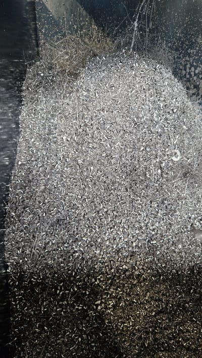 This image is a close-up, overhead shot of a pile of metallic shavings. The shavings are small, irregular in shape, and predominantly silver in color. They appear to be composed of aluminum or a similar metal, given their bright, reflective quality. Some of the shavings have a coiled or helical shape, suggesting they were produced by a machining process like drilling or milling.

The pile of shavings is substantial, filling a large portion of the frame. The texture is rough and granular, with many individual pieces visible. The lighting creates a play of light and shadow across the surface, highlighting the three-dimensionality of the pile. Towards the left side of the image, the shavings appear to be spilling onto a darker, smoother surface, which might be a metal tray or workbench. This darker surface shows some scratches and smudges, indicating it is a functional workspace. The background on the left is also darker and appears to be a metallic surface, possibly the side of a bin or machine.

There are no people visible in the image. The scene depicts the aftermath of a manufacturing or fabrication process, with the shavings representing waste material or byproducts. The overall impression is one of industrial activity and material processing. There is no indication of time of day or weather, and the lack of context beyond the visual suggests a purely functional or material study. No text is visible in the image. The location context of Ichenhausen, Germany, does not provide specific visual cues within the image itself.