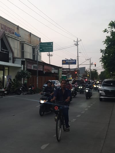 The image depicts a street scene in Proyonangg, Indonesia, likely in the late afternoon or early evening, given the soft, diffused light and the presence of headlights. A group of people on motorcycles and at least one bicycle are moving along a paved road. The road appears to be a main thoroughfare, with traffic lights and street signs visible.

In the foreground, a person is riding a bicycle, facing towards the camera. They are wearing dark clothing and appear to be looking at the camera. Behind them, several motorcycles are also visible, some with passengers. The motorcycles are illuminated by their headlights, adding to the low-light atmosphere.

Several buildings line the street, some of which appear to be shops or commercial establishments. There are trees and foliage visible, suggesting an urban or semi-urban environment. The sky is overcast, contributing to the muted colors of the scene.

A prominent green street sign is visible, indicating directions to "Semarang", "Kantor Bupati Batang", and "Pekalongan". Another sign reads "WILDHAN". The overall impression is one of everyday life, with people commuting or traveling on the road. The presence of multiple vehicles and pedestrians suggests a moderately busy area. There are no obvious signs of distress or unusual events; the scene appears to be calm and ordinary.