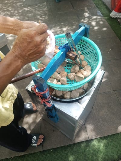 A vendor prepares food from a street cart. The cart, likely for boiling or deep frying, has a blue wooden frame holding a green plastic basket of rounded food items, possibly meatballs. The vendor is using tongs to pick food out of a plastic bag. He is wearing a yellow shirt, black pants, and sandals. The cart sits on a tile surface next to a patch of green grass.
