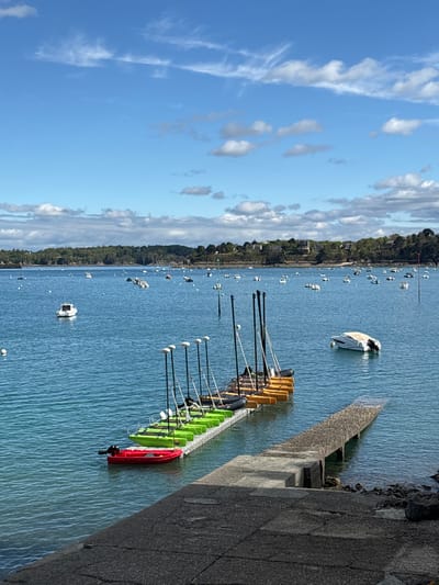 The photo shows a clear blue sky with white puffy clouds. In the foreground, a row of small green sailboats are docked along a floating dock.  The dock is connected to a concrete ramp leading up to a paved area. A red rowboat is docked at the end of the ramp.  There are many other boats in the distance scattered around the bay. The photo was taken in Dinard, France, at the coordinates 48.636, -2.049. The photo appears to be taken from a high vantage point overlooking the bay. The photo was likely taken in the late morning or early afternoon, as the shadows are not very long.