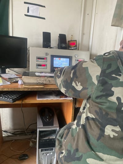 The image depicts an indoor scene within a facility in Dahiyat Harasta, Syria, focusing on a workstation. A person, whose back and right side are visible, wears a camouflage-patterned long-sleeved shirt and is positioned at a desk. Their right arm extends towards a control panel.
On the wooden desk, from left to right, are a black LG computer monitor, several papers, a small dish or ashtray, and a stack of books, one displaying Arabic script. A black computer keyboard rests further left on the desk. Below the desk, a silver desktop computer tower is situated on a lower shelf, and a black computer mouse is on the floor beside it. Numerous cables are visible connecting the equipment.
Behind the monitor and books, a large, light-grey control panel unit is present. On top of this unit are two black computer speakers and a separate small red digital display indicating "43.8". The face of the control panel features multiple smaller digital displays, showing values such as "0" and "190.6", alongside a central color touch screen interface displaying graphical process information. The person's right hand is near this touch screen, suggesting interaction with the controls.
The background consists of a light-colored, possibly white, painted wall. A piece of paper, secured with black tape, is affixed to the wall above the monitor. The overall environment appears functional, with visible signs of use and minor disarray.