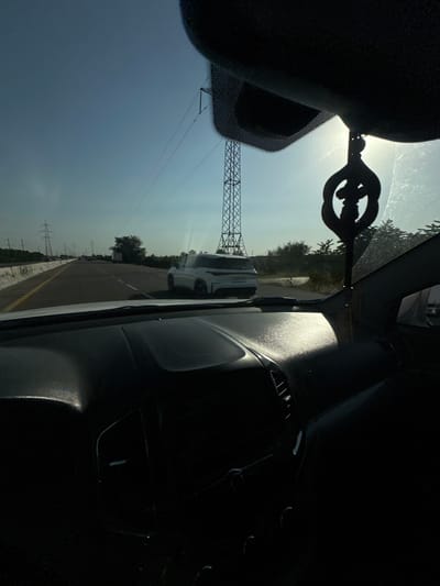 This media file captures a view from inside a car, looking out the windshield. The scene appears to be a road with a bright, hazy sky suggesting daytime, possibly late morning or early afternoon given the intensity of the light and shadows. The weather seems fair with clear skies, though there are some visible power lines and transmission towers suggesting an area with electrical infrastructure.
In the foreground, within the car's interior, a black, ornate object hangs from what appears to be a rearview mirror or similar fixture. It's a dark silhouette against the bright sky, with a distinctive circular design. The interior of the car is largely in shadow, with the dashboard and possibly a passenger seat visible.
Outside the car, a white vehicle is seen driving on the road, indicating that this is likely a highway or a main road. The road itself has a yellow line marking the lanes. On the sides of the road, there are trees and vegetation, and more power lines are visible stretching into the distance. The overall impression is of a road trip or daily commute through a landscape that combines natural elements with man-made infrastructure. The angle of the shot, looking slightly upwards and to the side, gives a dynamic perspective of the journey. There is no visible text in the image.