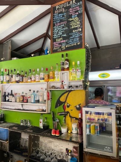 The image depicts a bar in Vagator, India, with a prominent blackboard menu listing popular drinks. The menu includes categories such as "Beer," "Whiskey," "Rum," "Vodka," and "Shots," with specific brands like Bud, Corona, Jameson, Jack Daniels, Old Monk, Bacardi, Rahasya, Absolute, and Grey Goose. A variety of liquor bottles are neatly arranged on shelves behind the counter. In the background, a person with dark hair pulled back is seen standing near a refrigerator. The bar area features a green painted wall, a cooler, and numerous glasses. A caricature of a yellow character, possibly Pikachu, is painted on the wall. The overall scene suggests a lively and well-stocked establishment.