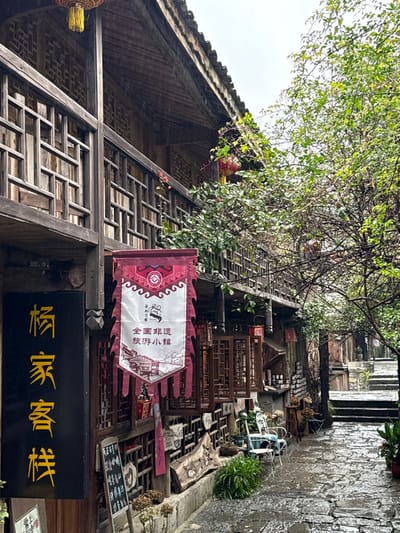 The image displays a narrow, wet, stone-paved alleyway in Gongtan, China, under an overcast sky. Traditional multi-story wooden buildings with dark facades, intricate wooden lattice balconies, and windows line both sides of the path.

On the left, a prominent vertical black sign with gold Chinese characters reading "杨家客栈" (Yang Family Inn) is affixed to the building. Below this, a large, vertically oriented red and white banner hangs, featuring a central white panel with a line drawing of traditional architecture and Chinese characters identifying the location as a "全国非遗旅游小镇" (National Intangible Cultural Heritage Tourist Town). A small chalkboard sign and decorative dried plants in pots are positioned at the base of this building, alongside a carved wooden log.

Further along the left side of the alley, two white metal chairs and a small round table with a light blue top are set on the wet cobblestone pavement, facing the street. Various potted plants and additional decorative objects are arranged near the buildings. Several red traditional Chinese lanterns and one yellow lantern are visible hanging from the eaves and balconies of the structures.

Abundant green foliage from trees and shrubs is present throughout the scene, with some branches overhanging the alley and others growing along the buildings. The stone pavement, reflecting light, indicates it is wet from rain or recent precipitation. The alley gently ascends into the background, where it narrows and transitions to stone steps. No individuals or animals are visible within the frame.