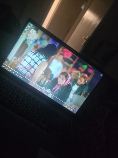 The image displays a laptop screen actively playing a video in a dimly lit indoor environment. No specific features identifying a location within Kaiserslautern, Germany, are discernible.
**Subjects/Objects:**
The primary subject is a laptop computer, with its screen prominently featured, showing a scene from a video. The laptop's keyboard is visible in the lower foreground. The video content includes multiple individuals. A person with long, dark hair wearing a white and black checkered or plaid top is seen from the back or side, facing away from the viewer. To the lower right of this person, a woman wearing a pink head covering (possibly a scarf or hijab) is visible, looking towards the right side of the screen. Other blurred figures and colorful decorations, resembling pompoms or balloons, are present in the background of the video. A 'B' logo is visible in the upper left corner of the video playback. A playback progress bar at the bottom of the screen indicates a duration of "03:15 / 07:02", alongside partially visible playback controls (play/pause, volume, settings, full screen icons). In the physical background of the room, a light-colored door with a dark handle is visible to the upper right of the laptop screen. A dark, possibly textured surface, consistent with bedding or fabric, is visible to the right of the laptop.
**Environment:**
The overall scene is dimly lit, suggesting low ambient light in the room. The primary light source appears to be the laptop screen itself. The door in the background suggests an interior residential or office space.
**Actions/Interactions:**
The laptop is engaged in playing video content. The individuals depicted in the video appear to be within a social setting, possibly interacting or observing.