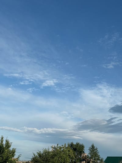 The media file shows a bright, clear blue sky with scattered, wispy white clouds. The image is oriented vertically, with the sky occupying the majority of the frame. On the right side of the image, a section of lush green trees is visible, with some branches extending into the lower part of the frame. Above the trees, there is a glimpse of a structure with a green, corrugated roof and wooden posts, suggesting a built environment. A thin, light-colored power line is also visible running through the trees. The clouds are more dense and elongated towards the right side of the sky, appearing somewhat hazy. The overall impression is a sunny day with pleasant weather, likely mid-morning or early afternoon given the brightness of the sky. No people or specific activities are depicted, making it a landscape-focused image. The location context is Tobyl, Kazakhstan.