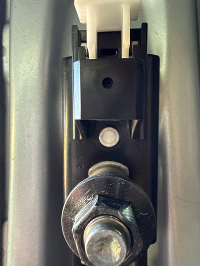 This is a close-up, eye-level shot of a mechanical component, likely part of a vehicle or machinery, seen in an indoor setting. The main subject is a black plastic housing with a white electrical connector attached to its left side. The connector has multiple pins visible.
A silver-colored bolt with a nut secures the black housing to a larger, metallic surface, which appears to be a panel or frame. The bolt head has hex-shaped edges, and there are scuff marks and some dirt on the bolt and nut, indicating use. A small, round, silver-colored screw is also visible near the center of the black housing.
The background is a textured, metallic surface that is mostly out of focus but appears to be a light gray or silver color. The lighting is somewhat harsh, creating highlights and shadows that emphasize the textures and details of the components. There are no people or animals present, and no discernible environmental cues to suggest the time of day or weather.
The image is focused on the mechanical details of the assembly. There is no clear indication of an activity taking place, but the presence of electrical connectors and bolts suggests it is part of a functional system. The overall impression is one of mechanical engineering and repair, possibly within a garage or workshop.
Faintly visible on the black housing, under the bolt and nut, are some numbers or markings. The clearest part of this text appears to be "2N3". The location context provided is Mastaga, Azerbaijan.
