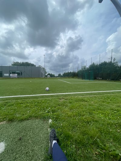 An outdoor shot depicts a soccer field with a low-angle perspective, looking across the green turf. In the foreground, a person's left leg, clad in dark blue pants and a black athletic shoe, is visible. The field is marked with white lines. A soccer ball rests on the grass near the center line. In the background, a grey building with a large sign displaying images of trucks is situated to the left. Beyond the building, a chain-link fence encloses the field, with trees and foliage visible behind it. The sky is overcast with dramatic, dark clouds. A lamppost stands tall near the building, and a soccer goal with a green net is visible on the right side of the field. The overall scene suggests a cloudy day at a sports facility. The location is identified as Jakai, Lithuania.