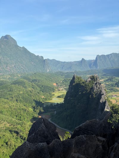 This is a wide-angle, eye-level shot of a lush, mountainous landscape under a clear blue sky. In the foreground, dark, jagged rock formations dominate the bottom of the frame, creating a dramatic natural frame. Beyond these rocks, the landscape unfolds into a series of rolling, verdant green hills covered in dense vegetation. A winding road cuts through the valley, bordered by patches of cultivated land, appearing as yellow and green rectangles, likely rice paddies. In the middle ground, a strikingly sharp, tree-covered rock peak rises dramatically, with what appears to be small structures or ruins on its summit. Further in the background, a range of majestic, mist-shrouded mountains extends to the horizon, their peaks softened by distance. The scene conveys a sense of remote, natural beauty, likely in a rural mountainous region such as Naka, Laos, where such dramatic karst topography is common. No people or animals are visible in the frame.
