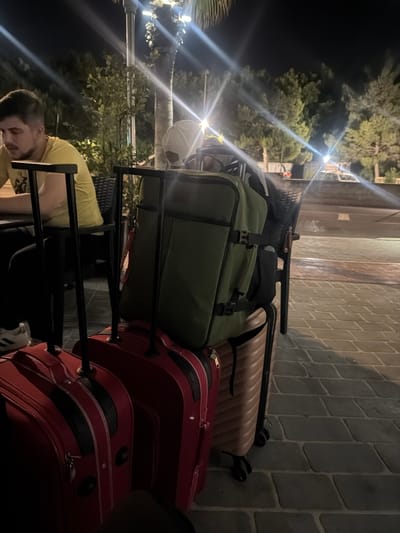 The image shows a stack of luggage on a luggage cart in Saint Paul's Bay, Malta. At the base of the cart is a red suitcase. Stacked on top of that, there is a second red suitcase. On top of that is a green bag. A tan suitcase is located on the top. A person wearing a yellow shirt is holding the cart with the luggage. A person wearing a white hat is next to the cart. The background shows the outdoor setting, which appears to be near a sidewalk or street. Trees and streetlights are visible. The photo appears to have been taken at night or in a dimly lit environment.