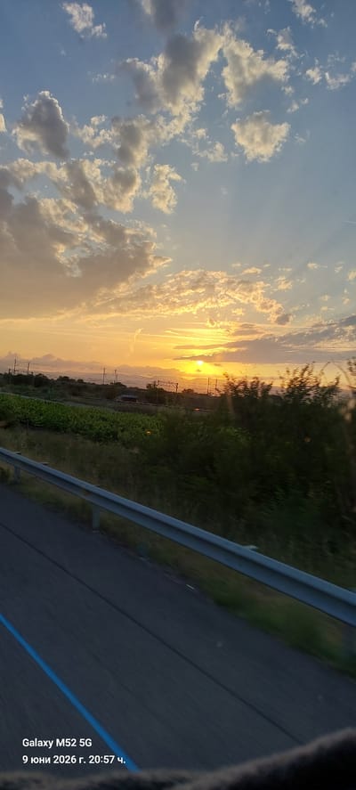 This is a photograph taken from a vehicle, likely a car or bus, showing a landscape during sunset. The sky is filled with dramatic clouds, illuminated by the setting sun which is a bright orange circle just above the horizon. The sun's rays create a warm, golden glow across the sky. In the middle ground, there is a stretch of green vegetation, possibly fields or vineyards, with some trees and what appear to be utility poles and wires. A road with a guardrail is visible in the foreground, suggesting the perspective is from a moving vehicle. The bottom left corner of the image contains a timestamp and device information: "Galaxy M52 5G 9 юни 2026 г. 20:57 ч.". The location provided, Can Cartró, Spain, is a small village or hamlet. The scene depicts a rural or semi-rural environment at dusk. There are no people or discernible actions.