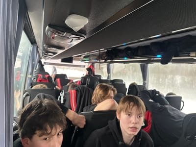 The image captures the interior of what appears to be a passenger bus or coach, likely during a journey. The perspective is from within the bus, looking towards the front and slightly to the right side.
Several young people, primarily teenagers or pre-teens, are visible as passengers. In the bottom left corner, part of a person's face with brown, medium-length hair is visible, looking towards the camera. More prominently in the bottom right, another young person with light brown, straight hair and wearing a black hoodie is looking slightly to the right, their expression appearing calm or neutral. Further into the bus, other passengers are seen in various states of relaxation; one person with blondish hair is visible from the back of their head on the left, while another's legs, clad in grey socks, are propped up on a seat in the middle-ground. Many of the passengers are wearing dark clothing, including black hoodies or jackets, some with red accents or stripes on their backpacks or clothing.
The bus interior features dark-colored, presumably fabric or leather, seats with high backs and headrests. Above the seats, dark gray overhead compartments are present, beneath which small blue lights are visible. Individual overhead control panels, containing reading lights and air vents, can be seen along the ceiling. A white, somewhat conical object is attached to the ceiling closer to the front of the bus. Large windows line the left side of the bus, revealing a wintry landscape outside, characterized by snow and trees, suggesting cold weather conditions. The light filtering through the windows indicates it is daytime, possibly overcast. Several bags and backpacks, some with red elements, are visible on or around the seats, indicative of travel luggage.
The overall impression is that of a group of young people traveling together on a bus through a snowy environment. No discernible text is visible within the image.