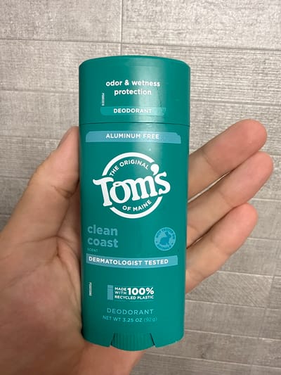 An overhead, close-up shot shows a hand holding a teal-colored stick of Tom's of Maine "Clean Coast" deodorant against a grey, textured background. The deodorant stick has white text that reads "odor & wetness protection," "DEODORANT," "ALUMINUM FREE," "THE ORIGINAL TOM'S OF MAINE," "clean coast SCENT," "DERMATOLOGIST TESTED," and "MADE WITH 100% RECYCLED PLASTIC." A circular emblem with a bunny inside that reads "NOT TESTED ON ANIMALS" is also visible. The hand is positioned in the lower left and right of the frame, obscuring part of the deodorant stick. The image appears to have been taken indoors. The setting is not identifiable as Burbank, United States.