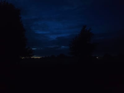 The image captures a dark, moody outdoor scene, likely at dusk or twilight, in the Яглевіцкі сельскі Савет, Belarus. The sky is a deep, ominous blue with wispy, illuminated clouds that suggest residual light from the setting sun. Silhouetted against the sky are the dark, indistinct shapes of trees and vegetation, creating a somber and somewhat melancholic atmosphere. The foreground is almost entirely black, indicating the absence of significant light sources and obscuring any details of the ground or any potential structures. The overall impression is one of a quiet, still evening in a rural or semi-rural setting.