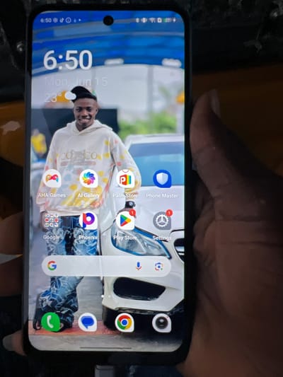A black smartphone displays a lock screen with a time of "6:50" and the date "Mon, Jun 15" with a temperature of 23 degrees Celsius. The background features a young Black man wearing a white hooded sweatshirt with a colorful tie-dye pattern and a black beanie. He is smiling and looking at the camera. The lower portion of the screen shows various app icons including AHA Games, AI Gallery, Palm Store, Phone Master, Google, Phoenix, and Play Store. A Google search bar with a microphone icon is also visible. The phone is held by a person whose hand is partially in the frame on the right side of the image. The background appears to be outdoors with a car partially visible. The image quality is moderate. There is no specific information to indicate the location in the city of Orifite, Nigeria.