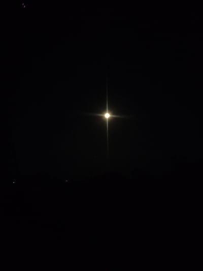 The image depicts a night scene, likely taken outdoors in Kostiantynivka, Ukraine. The dominant subject is a very bright, artificial light source in the center of the frame, casting a strong glare and a distinct starburst effect. This light is likely a street lamp or a similar overhead fixture. The background is almost entirely black, indicating nighttime. There are faint hints of distant lights or structures along the bottom horizon, suggesting a populated area, possibly a town or city.
In the upper left corner, there are a few small, indistinct purple or pinkish shapes, possibly reflections or artifacts. Two very small, faint dots of light are visible in the lower left quadrant. The overall impression is one of darkness with a single, powerful light source dominating the scene. There is no clear indication of weather conditions, but the darkness implies it is not daytime. The scene appears still and quiet, with no visible people or active events. The focus is on the stark contrast between the intense light and the surrounding darkness.