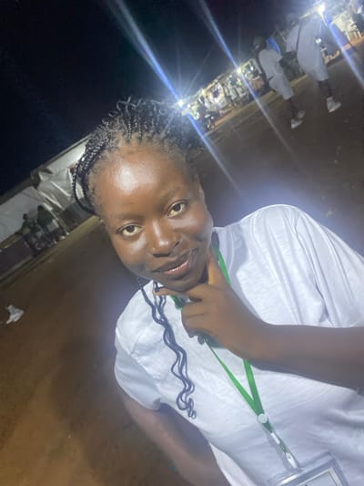 A medium-shot, low-angle, eye-level, indoor and outdoor, night time, slightly overexposed photograph shows a young woman with dark skin, dark eyes, and braided dark hair posing for a selfie. She is wearing a white t-shirt with a green lanyard and a clear plastic badge. Her right hand is resting on her chin and neck. The background shows several people in white clothing in front of tents and stalls, illuminated by bright lights. The ground appears to be dirt or sand.