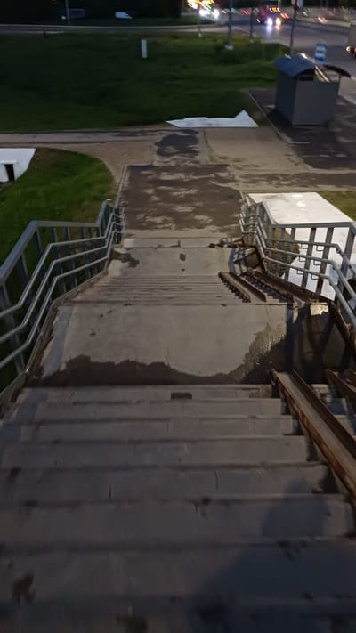 The image shows a concrete structure with a staircase and metal railings. The location appears to be in an urban area, likely in Obukhovo, Russia, based on the information provided. To the left of the structure, there is a grassy area with trees and streetlights visible in the distance, suggesting a street or park. The photo seems to have been taken during the evening or night, as indicated by the streetlights and darker lighting. The concrete appears to be damp or wet, indicating rain or recent cleaning. The scene is devoid of people.