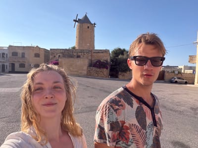 In Xagħra, Malta, a young blonde woman with fair skin and wavy blonde hair is taking a selfie with a young man in the foreground. The woman is wearing a light-colored top and is smiling slightly. The man, to her right, is wearing sunglasses and a t-shirt with a tropical leaf pattern. Behind them, a traditional stone windmill with a dark conical roof stands against a clear blue sky. The windmill is surrounded by stone buildings and some vegetation. The ground is paved with asphalt, and a silver car is parked in the background. The overall scene appears to be set in a historical part of the town on a sunny day.