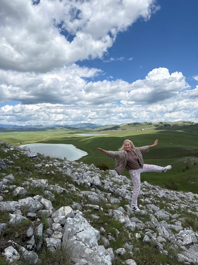 The image displays an adult female posing on a rocky hillside, set against a vast, verdant mountainous landscape under a partially cloudy sky. The location is identified as Javorje, Montenegro.

The woman, positioned slightly right of center in the mid-ground, has blonde hair and is smiling towards the camera. She is wearing a light brown quilted jacket over a solid brown t-shirt and light-colored plaid trousers. Her footwear consists of white athletic shoes with a black "N" logo on the side. She is captured mid-action, balancing on her left foot with her right leg extended horizontally to her side. Both arms are outstretched horizontally, palms open and facing forward.

The immediate foreground consists of numerous irregularly shaped grey and white rocks of varying sizes, interspersed with sparse green grass and some small white wildflowers. The terrain slopes downwards from right to left.

In the mid-ground and background, a wide expanse of rolling green hills and valleys dominates the scene. A light blue-green lake, roughly heart-shaped, is prominent on the left side of the mid-ground, with a light-colored dirt road winding along its edge and into the distance. Additional, smaller water bodies are faintly visible further into the landscape. Scattered, small, light-colored structures, resembling rural houses or farm buildings, are discernible on the distant green hills. The horizon is defined by undulating mountain ranges, also covered in green vegetation.

The sky occupies the upper portion of the image, featuring a mix of bright white cumulus and stratocumulus clouds against patches of clear blue sky. The lighting suggests bright daylight conditions.