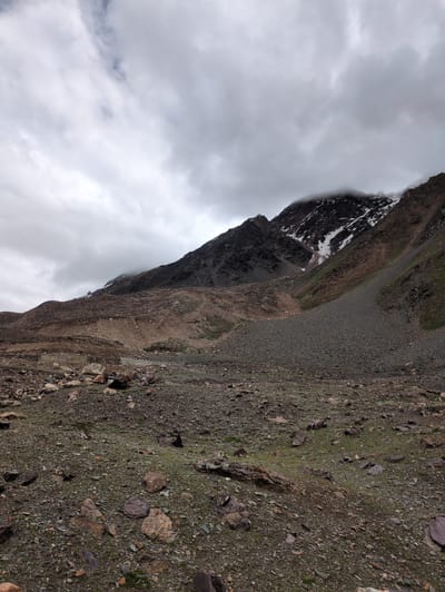 This image captures a desolate mountainous landscape in Zing Zing Bar, India. The foreground is a rocky, uneven terrain sparsely covered with small patches of green vegetation. Large and small stones litter the ground, varying in color from dark grey to reddish-brown. In the mid-ground, a steep, rocky slope rises, characterized by scree and loose rocks. Further back, the terrain continues to ascend, forming a dark, rugged mountain range. Patches of snow are visible on the higher slopes, and some areas are shrouded in thick, grey clouds that obscure the mountain peaks. The sky is overcast with heavy, diffused clouds, contributing to the somber and wild atmosphere of the scene. There are no people or man-made structures visible in the image.