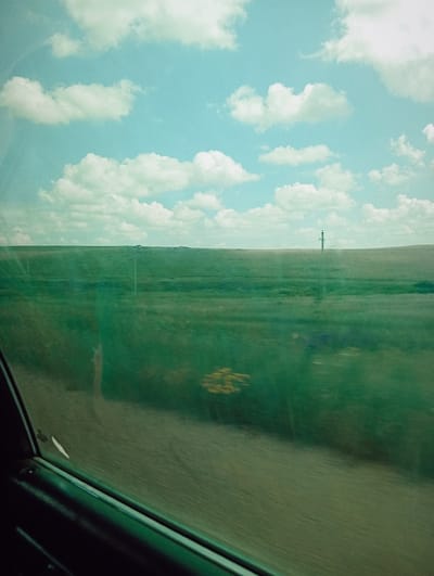 The image captures a view from inside a vehicle looking out a window. The scene depicts a vast, green, grassy plain under a bright blue sky with scattered white clouds. A single, tall, slender pole with horizontal attachments is visible in the distance on the horizon line. The foreground shows the interior of the vehicle, including a dark-colored window frame and the passenger side door panel. There are reflections on the window, suggesting the image was taken while in motion. The overall impression is of a rural or steppe landscape. The location is identified as Aleksandrovka, Kazakhstan.