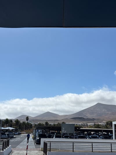 The image captures an outdoor scene, likely a parking area or a commercial complex, set against a dramatic backdrop of arid mountains under a bright sky. The perspective is from a slightly elevated vantage point, possibly a building balcony or upper level.

On the left side of the frame, a large, dark, metallic-clad structure or building facade extends into the foreground, occupying roughly a quarter of the image. The upper portion of the scene is dominated by a clear, vibrant blue sky. A long, well-defined band of white cumulus clouds stretches horizontally across the middle of the sky, creating a distinct line above the distant landscape.

In the mid-ground and background, a range of imposing, barren mountains with visible rocky textures and geological striations rise steeply. The peak of the closest mountain is prominent on the left side of the background, appearing dark and rugged. Below the mountains, there are buildings and clusters of palm trees, indicating a developed area.

To the right, a multi-level structure, possibly a parking garage or part of a building, features white walls and metallic railings. Beneath this structure and stretching across the lower right, numerous cars are parked, some under a covered canopy. A road or paved area is visible, traversed by a red and white striped barrier. One person, dressed in dark attire, is seen near this barrier, appearing to walk or stand. A dark rectangular board, possibly a bulletin board, covered with several small, light-colored notes or stickers, stands near the parked vehicles.

The scene is illuminated by bright daylight, suggesting a sunny day with clear weather, consistent with the reported location of San Bartolomé, Spain, known for its volcanic landscapes and palm-fringed areas. No legible text is discernible on any objects or signs within the image.