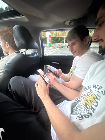 The image depicts the interior of a vehicle, likely parked or stopped at a gas station in Fort Lee, United States. Three male individuals are visible within the car.

The leftmost occupant, positioned in what appears to be the driver's seat, is seen from the back of the head and shoulder; he has curly brown hair and wears a light-colored shirt, facing forward.

In the rear passenger area, two males are present. The individual on the left has short dark hair and wears a white t-shirt with a green graphic design; he is looking down at a black smartphone held in both hands. The second individual, positioned to the right and partially obscured, shows a profile of his face with a beard and wears a white t-shirt with a visible graphic. His right hand holds a black smartphone. In the immediate foreground, a pair of hands, likely belonging to this second rear passenger, are seen actively interacting with a light-colored smartphone. These hands are positioned over black sweatpants featuring a white Nike logo.

Through the passenger window, a gas station environment is visible, including a multi-segment digital sign displaying prices in red and green, and part of a gas pump canopy structure. Green foliage is visible behind the gas station elements. A sunroof is partially open above the rear seats. All three individuals are focused on their respective smartphones.