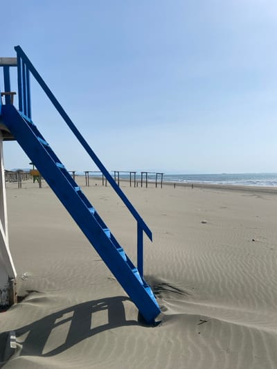 The image displays a sunlit, sandy beach located in Ulcinj, Montenegro. In the foreground, a prominent bright blue wooden staircase, equipped with a handrail, ascends diagonally from the lower left to the upper left of the frame. Its base is partially enveloped by light grey sand, which exhibits wind-formed ripple patterns. A sharp, elongated shadow of the staircase is cast across the sand. A portion of a white vertical support structure is visible on the far left, indicating the staircase is part of a larger, unseen platform.

The midground features numerous skeletal wooden structures, consisting of vertical poles and horizontal crossbeams, sparsely distributed across the sand toward the water. Some structures show remnants of netting or fabric. The background reveals a calm, light blue-grey sea with a discernible horizon. A dark, elongated object, possibly a distant vessel, is visible on the water near the horizon line. The sky above is clear and light blue. The beach is devoid of people, and minor pieces of debris are scattered across the sand.