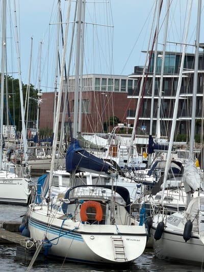 The image depicts a harbor scene in Wismar, Germany, with numerous sailboats docked. The foreground features several white sailboats, one prominently displaying the name "Wismar" on its stern, indicating its origin or affiliation with the city. These sailboats are tightly packed, with their masts and rigging creating a dense network of lines against the overcast sky. The boats are moored to a wooden dock. In the background, modern brick buildings and apartments with dark balconies and glass facades are visible, some with trees and greenery interspersed. The water in the harbor appears calm. No people are visible in the image.